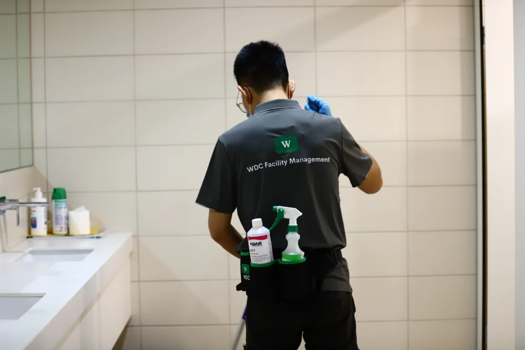 Rear view of a cleaner wearing a backpack vacuum in a tiled commercial restroom.