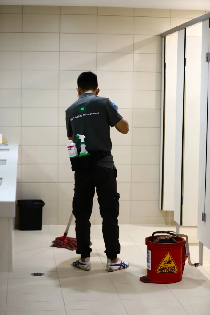 Uniformed janitorial staff member carrying vacuum equipment through a facility hallway.