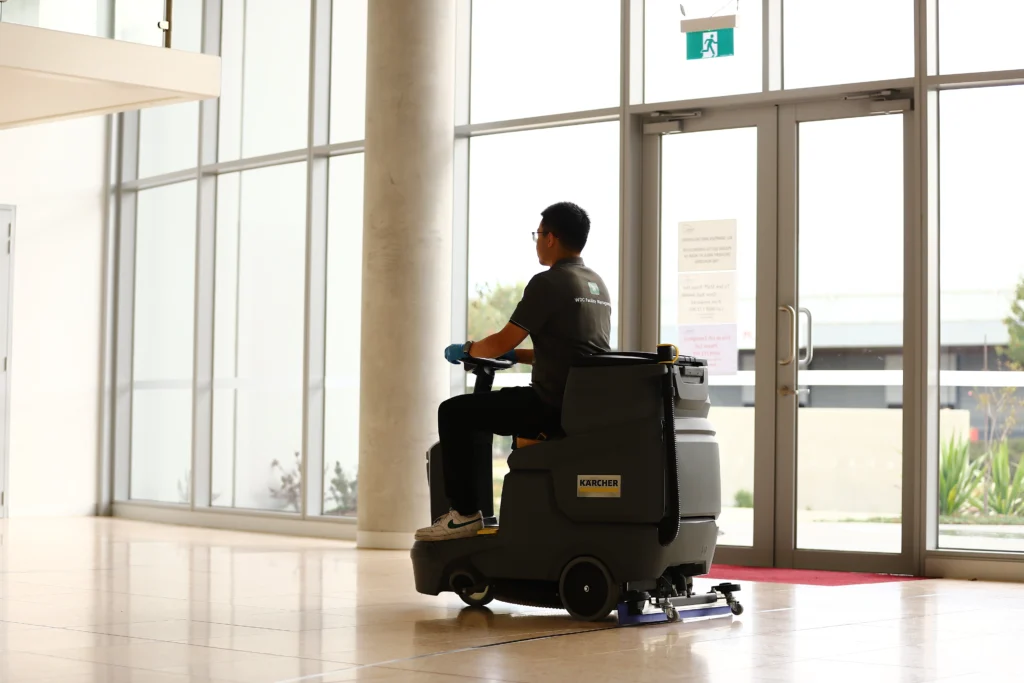Operator driving a ride-on floor scrubber in a spacious sunlit building lobby.