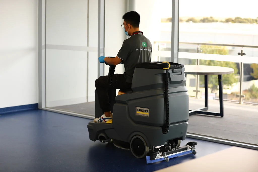 Ride-on floor scrubber washing hard floors in a large commercial office corridor.