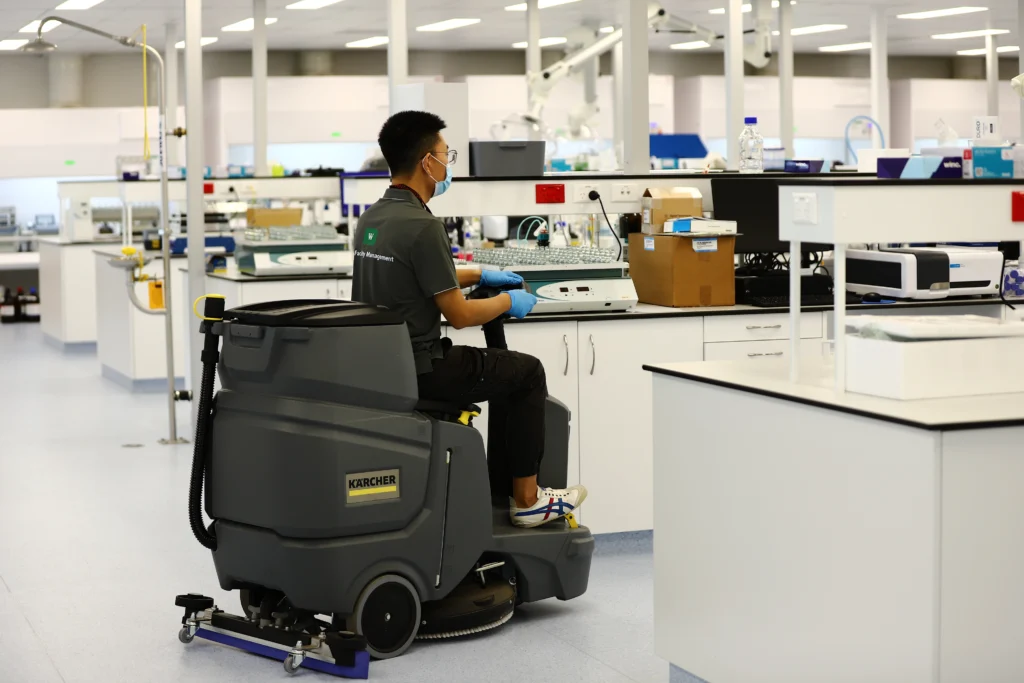 Commercial cleaning technician operating a ride-on floor scrubber in a sterile laboratory environment.