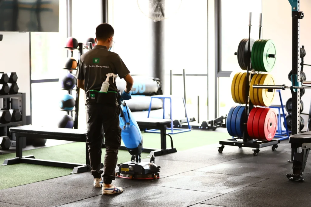 Cleaner mopping rubber gym flooring near weight racks in a commercial fitness center.
